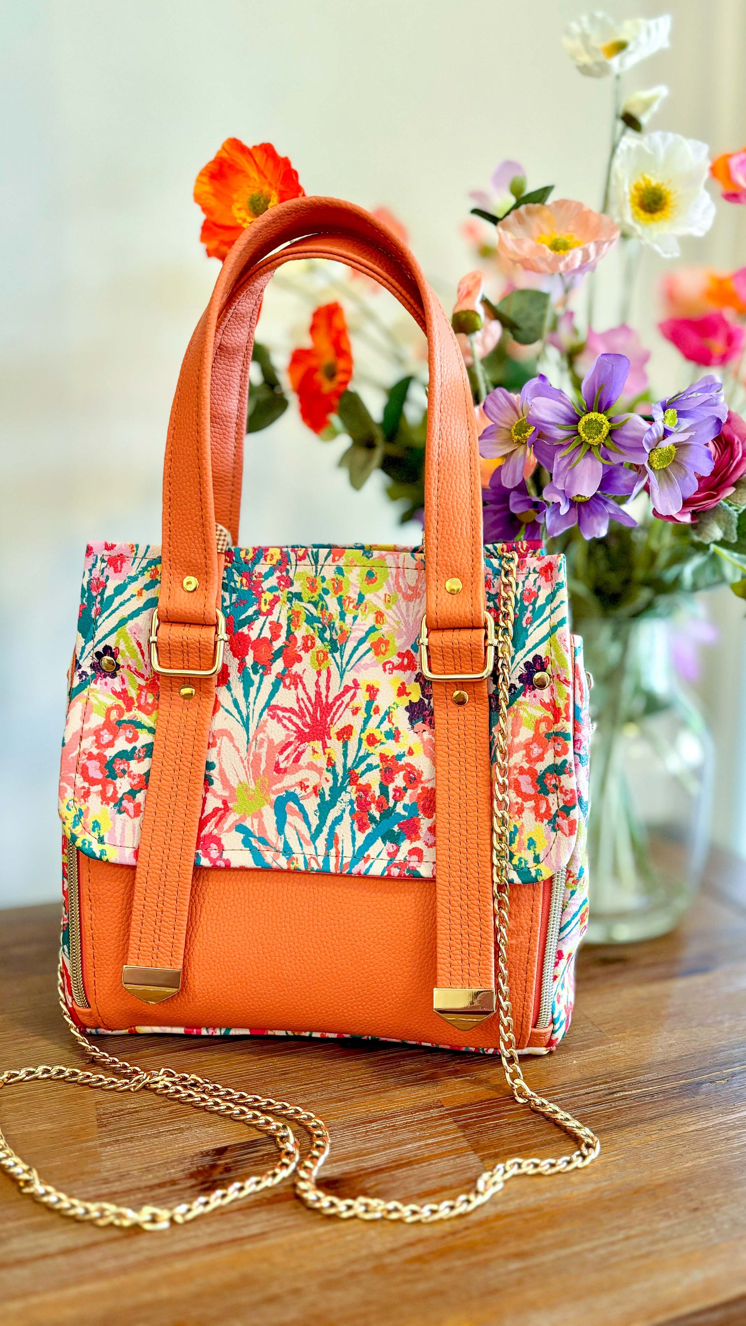 Colorful floral handbag with orange accents on a wooden surface, with flowers in the background.