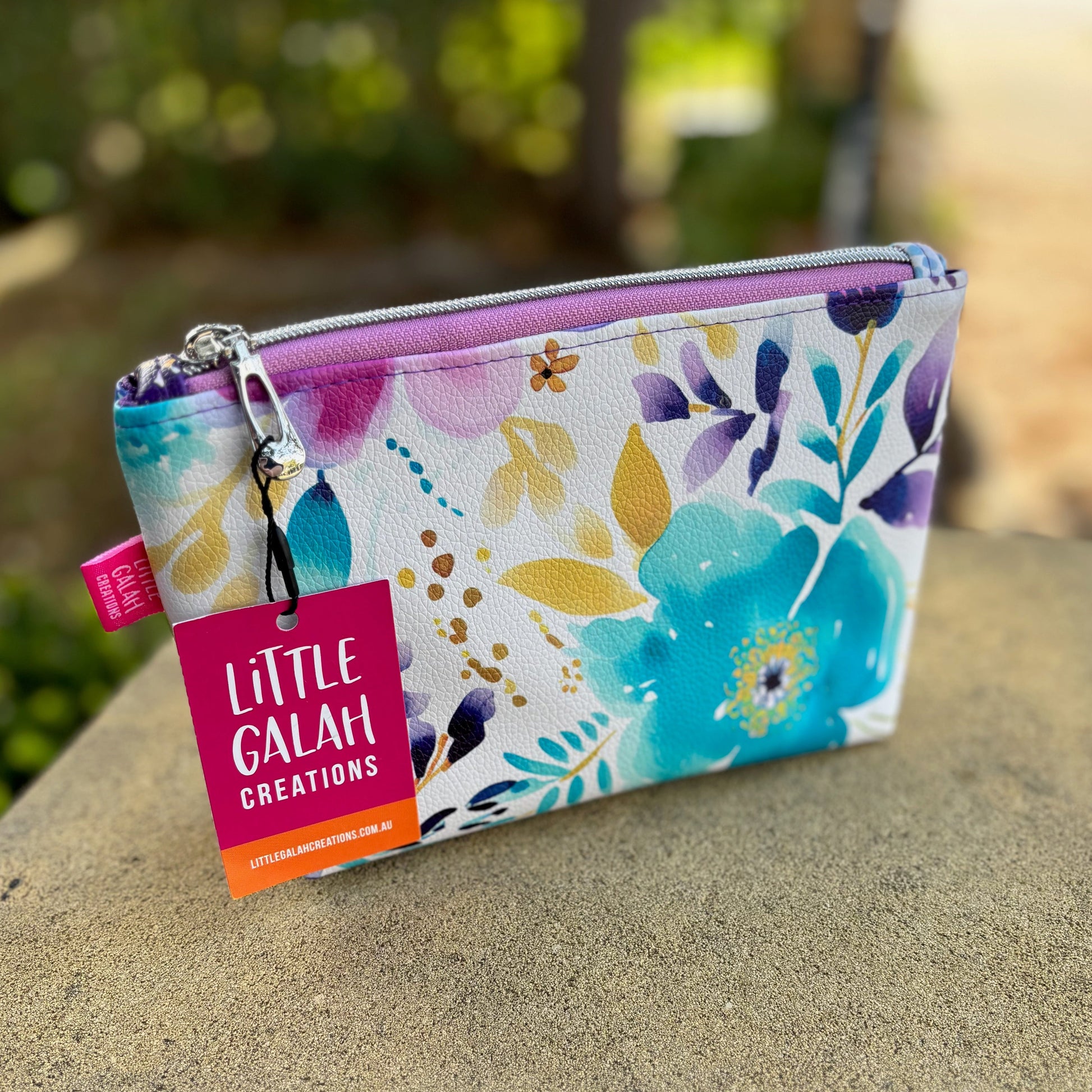 Floral pouch with 'Little Galah Creations' tag on a stone surface outdoors