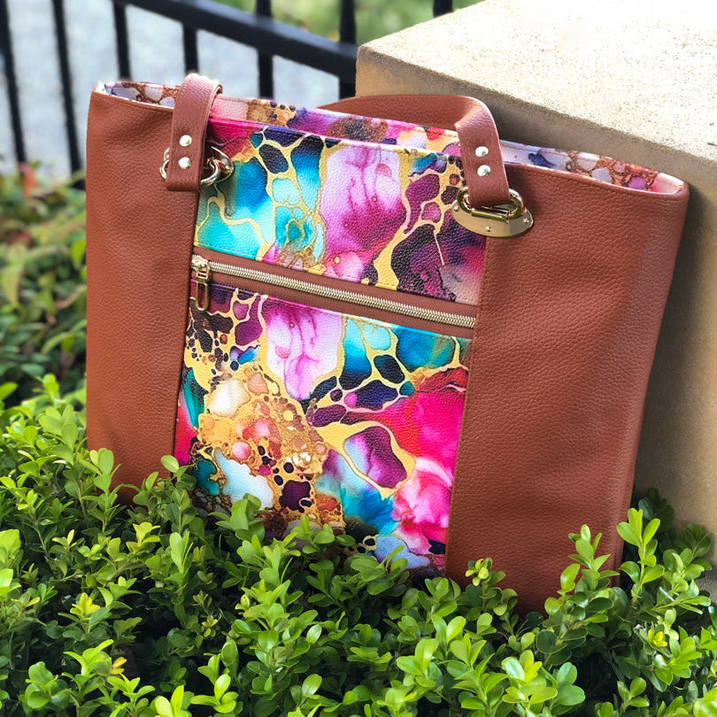 Multicolored floral-patterned handbag with brown leather accents on a green bush.