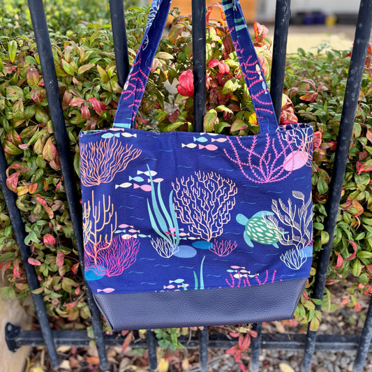 Blue tote bag with coral reef design hanging on a black metal fence.
